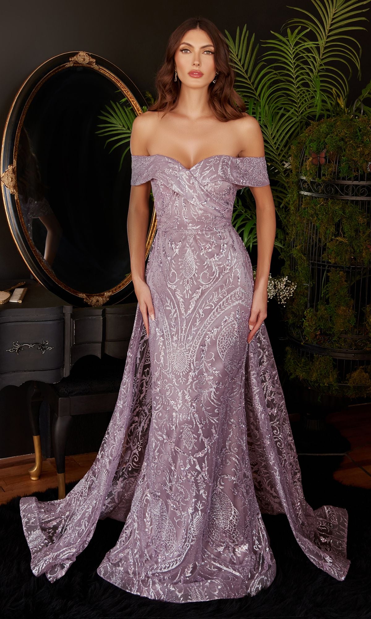 A woman stands indoors in the Long Prom Dress with Glitter-Print Overskirt J836, featuring a lilac off-the-shoulder style, baroque glitter prints, and a flowing train. A round mirror, black vanity, and green plants decorate the background.